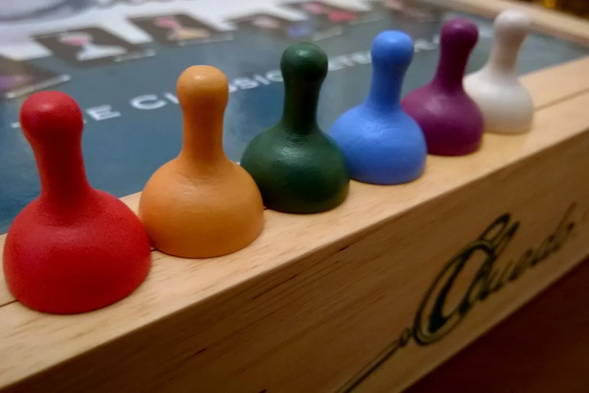 Six coloured suspect pawns lined up along the wooden edge of a Cluedo set, in red, orange, green, blue, purple and white